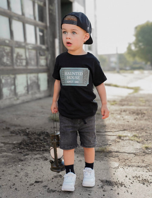 A toddler boy wearing a black graphic t-shirt with 'Haunted House' printed on the front, gray shorts, white sneakers, and a black baseball cap, standing outdoors on a cracked concrete ground next to a building.