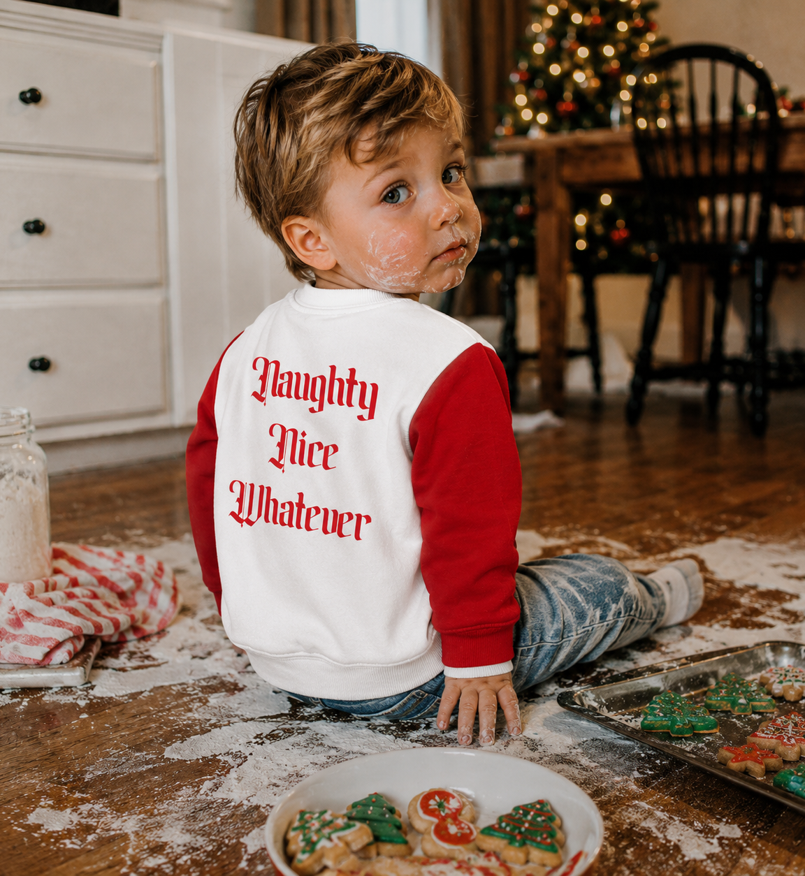 A young child sitting on a wooden floor covered in flour, wearing a red and white long-sleeve shirt with the phrase 'Naughty Nice Whatever' printed in red gothic letters on the back. The child has flour on their face, with decorated Christmas cookies on a tray and a plate nearby, and a decorated Christmas tree in the background.