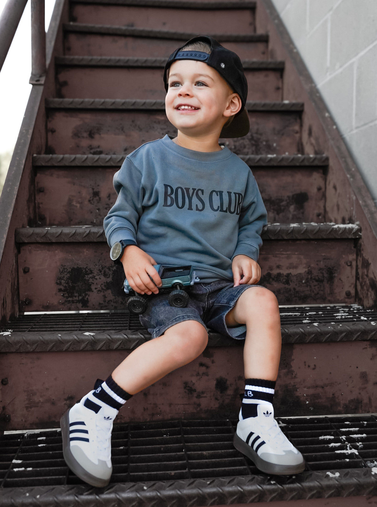 A young boy sitting on an outdoor metal staircase, wearing a slate blue long-sleeve sweatshirt with 'BOYS CLUB' printed across the chest, denim shorts, white sneakers, and a black baseball cap.