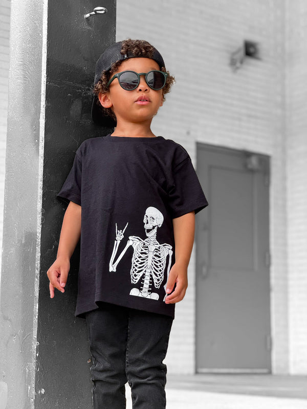 Child wearing a black t-shirt with a skeleton design, sunglasses, and cap against a gray building background.