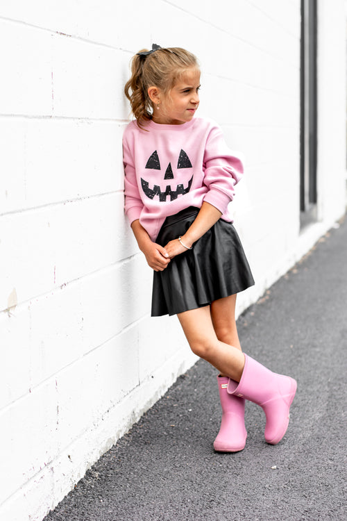 Child wearing a pink sweater with a Halloween design, black skirt, and pink boots against a white wall.