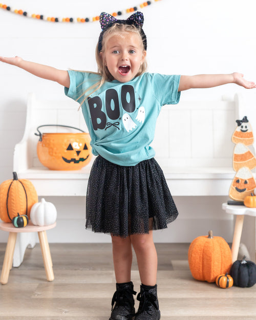 A young girl with blonde hair in a black cat-ear headband stands with arms outstretched, wearing a light blue graphic t-shirt that says 'BOO' in black letters with white ghosts, a black tulle skirt, and black sneakers. She is posing in a decorated Halloween room with pumpkins, a jack-o'-lantern bucket, and a candy corn decoration.