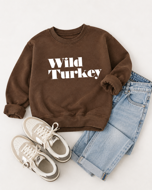 A brown crewneck sweatshirt with 'Wild Turkey' printed in white across the chest, styled in a flat lay with light blue jeans and beige sneakers on a light off-white background.